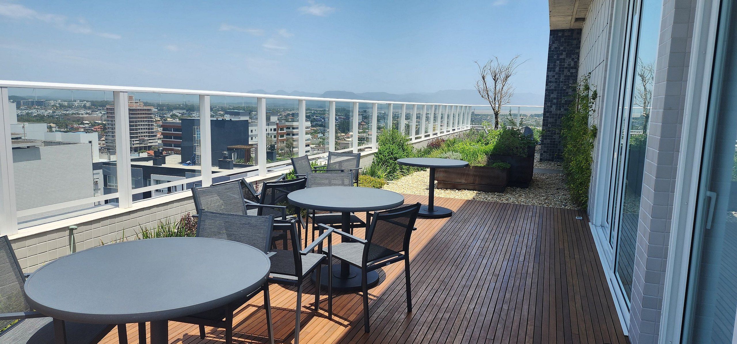 A rooftop terrace with two round tables and chairs, a glass railing, and a view of the cityscape.
