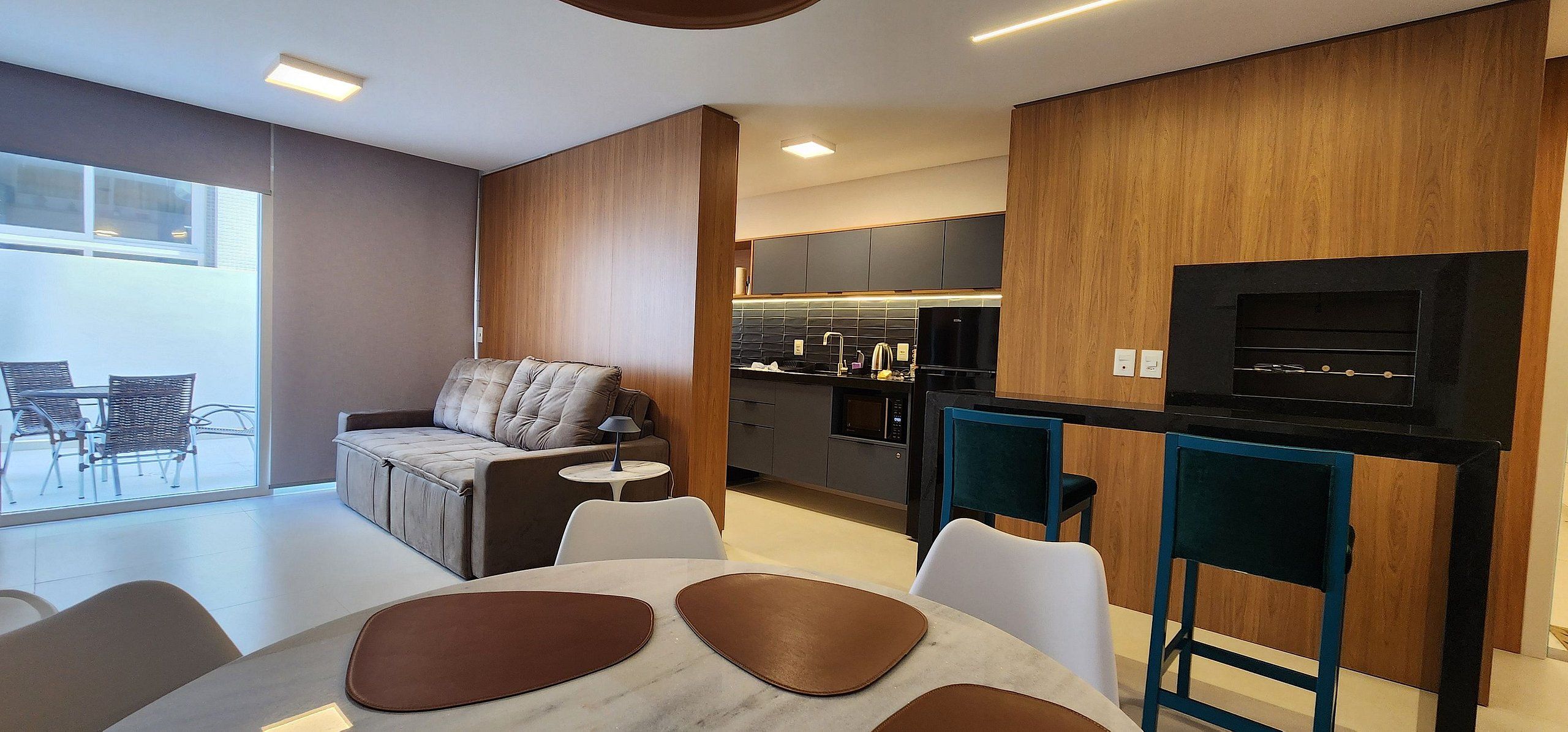 A modern kitchen with a built-in oven, microwave, and black TV. There is a dining area with a table and chairs, and a sofa with a lamp.