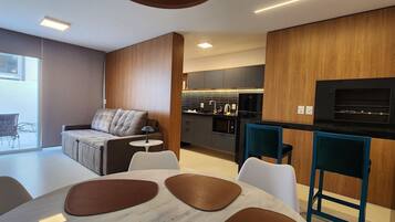 A modern kitchen with a built-in oven, microwave, and black TV. There is a dining area with a table and chairs, and a sofa with a lamp.