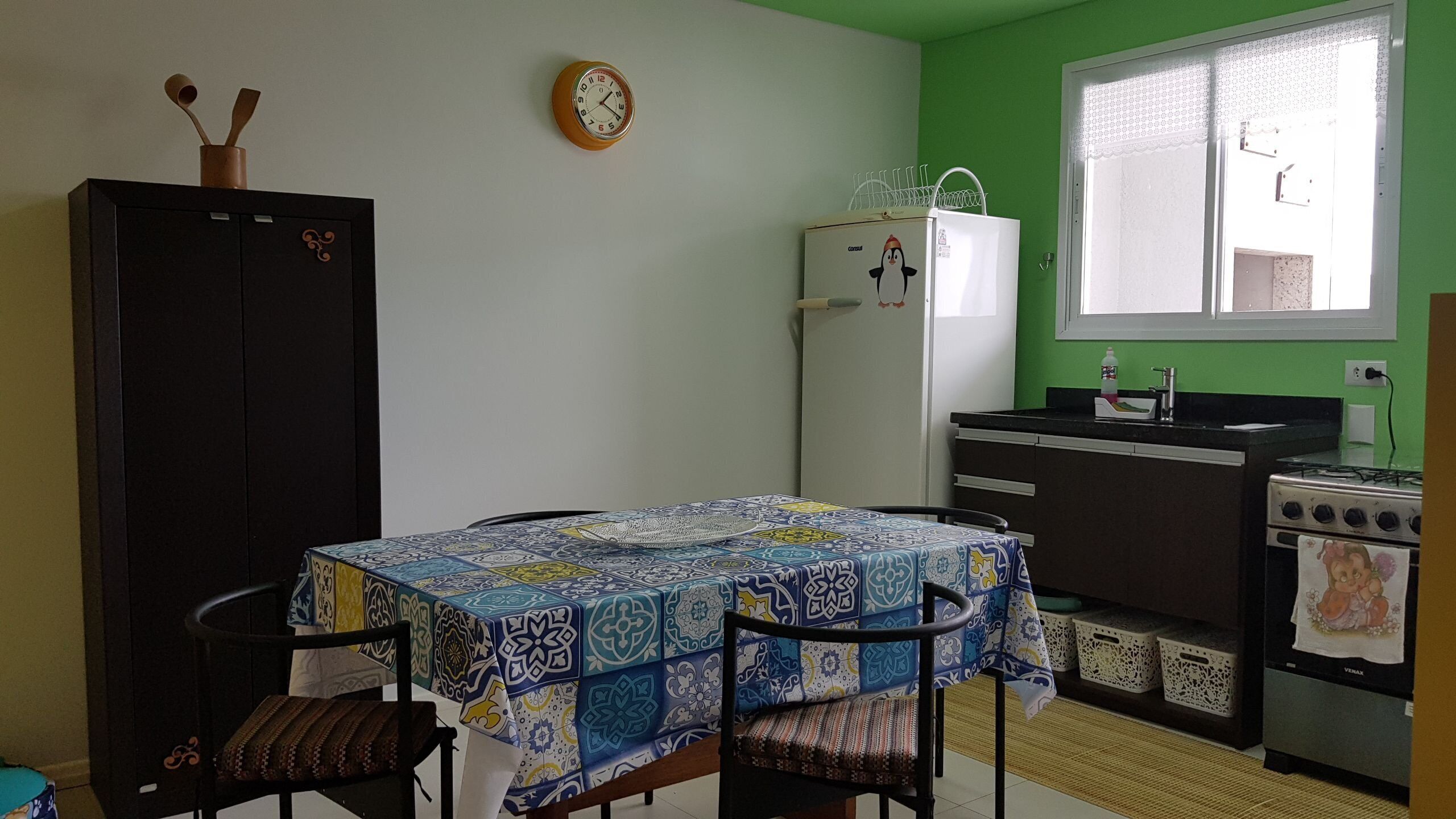 A compact kitchen with a dining table, chairs, a refrigerator, a stove, and a clock on the wall.
