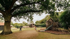 Een speeltuin met een glijbaan en klimconstructies, omgeven door bomen en een picknickplaats.