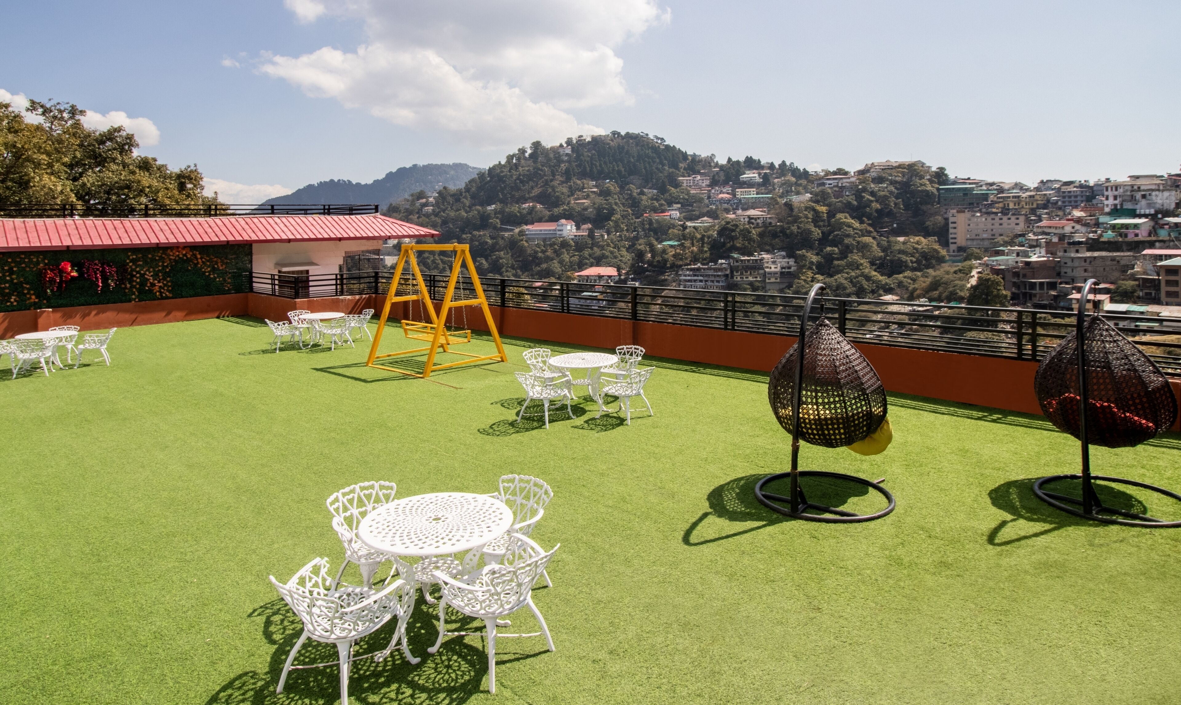 Une terrasse sur le toit avec un revêtement de gazon synthétique, des tables et des chaises en métal blanc, une balançoire jaune et des fauteuils suspendus.