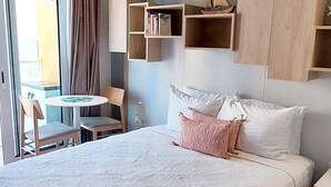 Une chambre d’hôtel avec un grand lit, une petite table ronde et une chaise. Au-dessus du lit, on trouve des étagères avec des livres et un petit modèle de bateau.