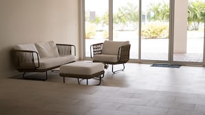 Une salle d’attente avec deux fauteuils en osier et un canapé, un sol carrelé et une vue sur les palmiers à travers de grandes fenêtres.