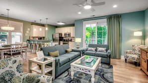 A living room with a sofa, coffee table, and patterned chairs. There is a dining area with a table and chairs, and a kitchen area with cabinets and appliances.