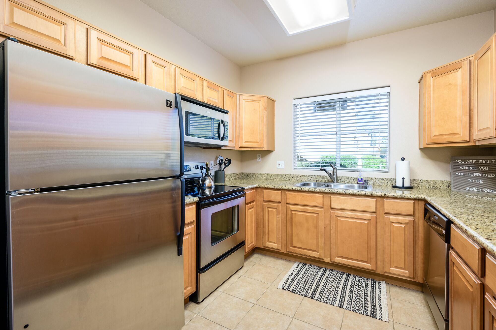 A kitchen with wooden cabinets, a stainless steel refrigerator, a microwave, an oven, a sink, and a window with blinds.