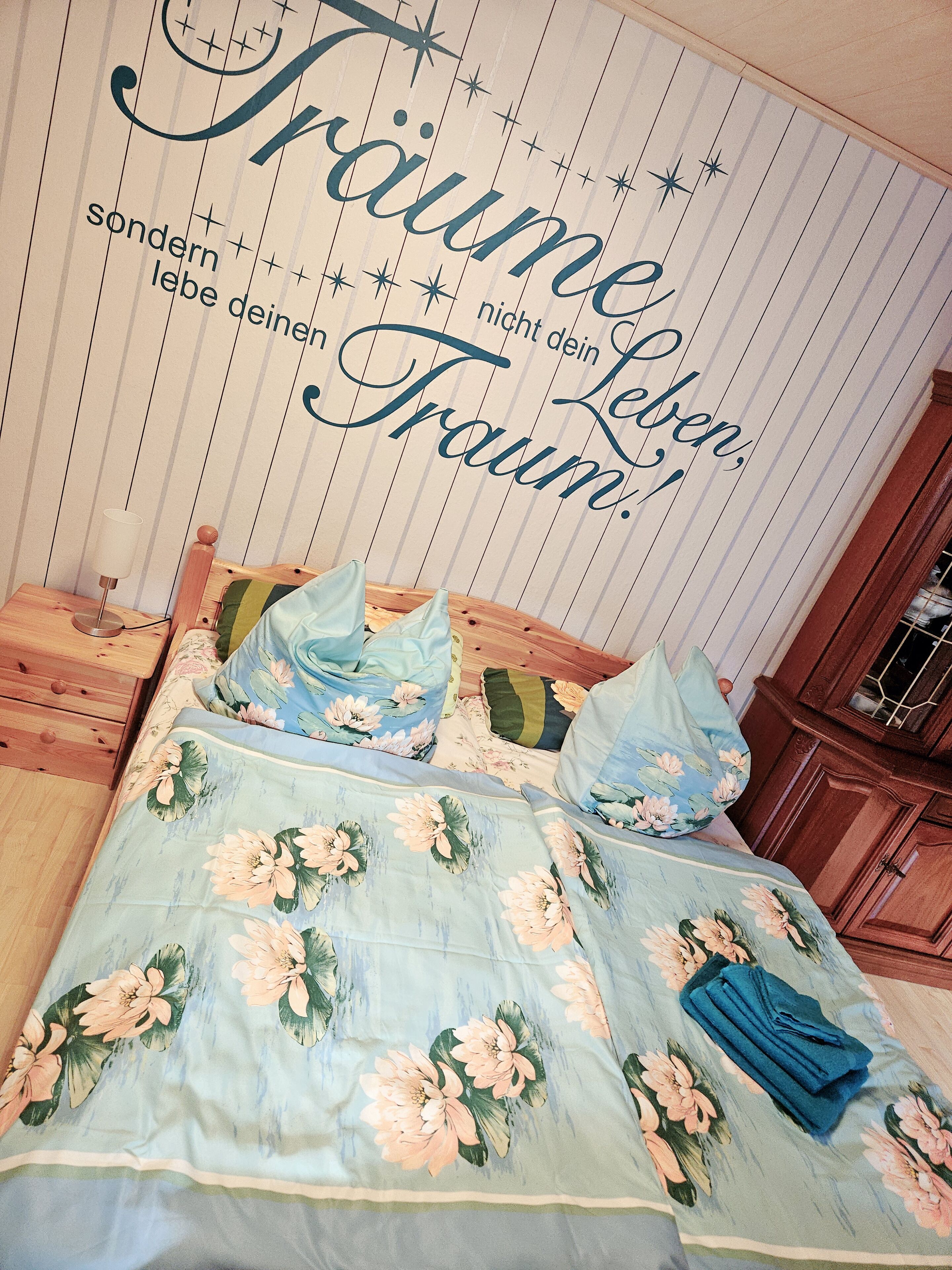 Quarto com cama decorada com roupa de cama floral, luminária de cabeceira e parede com texto decorativo.