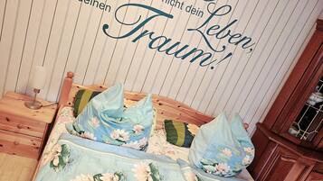 Quarto com cama decorada com roupa de cama floral, luminária de cabeceira e parede com texto decorativo.
