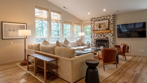 A spacious living room with a large sofa, a coffee table, and a fireplace. There are multiple windows providing natural light, and a television mounted on the wall.