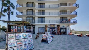 Un edificio de varios pisos con balcones, una tienda con un letrero que dice 'TIKI' y 'PIZZA', y una exhibición de obras de arte coloridas en primer plano.