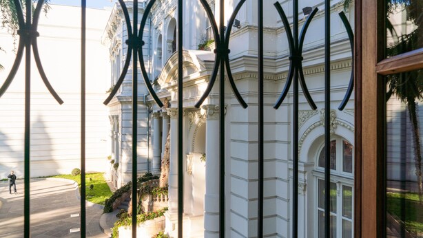 A view through a metal gate of a classical building with columns and a person walking on the street. - H&B apartments Atoneli (Tbilisi)