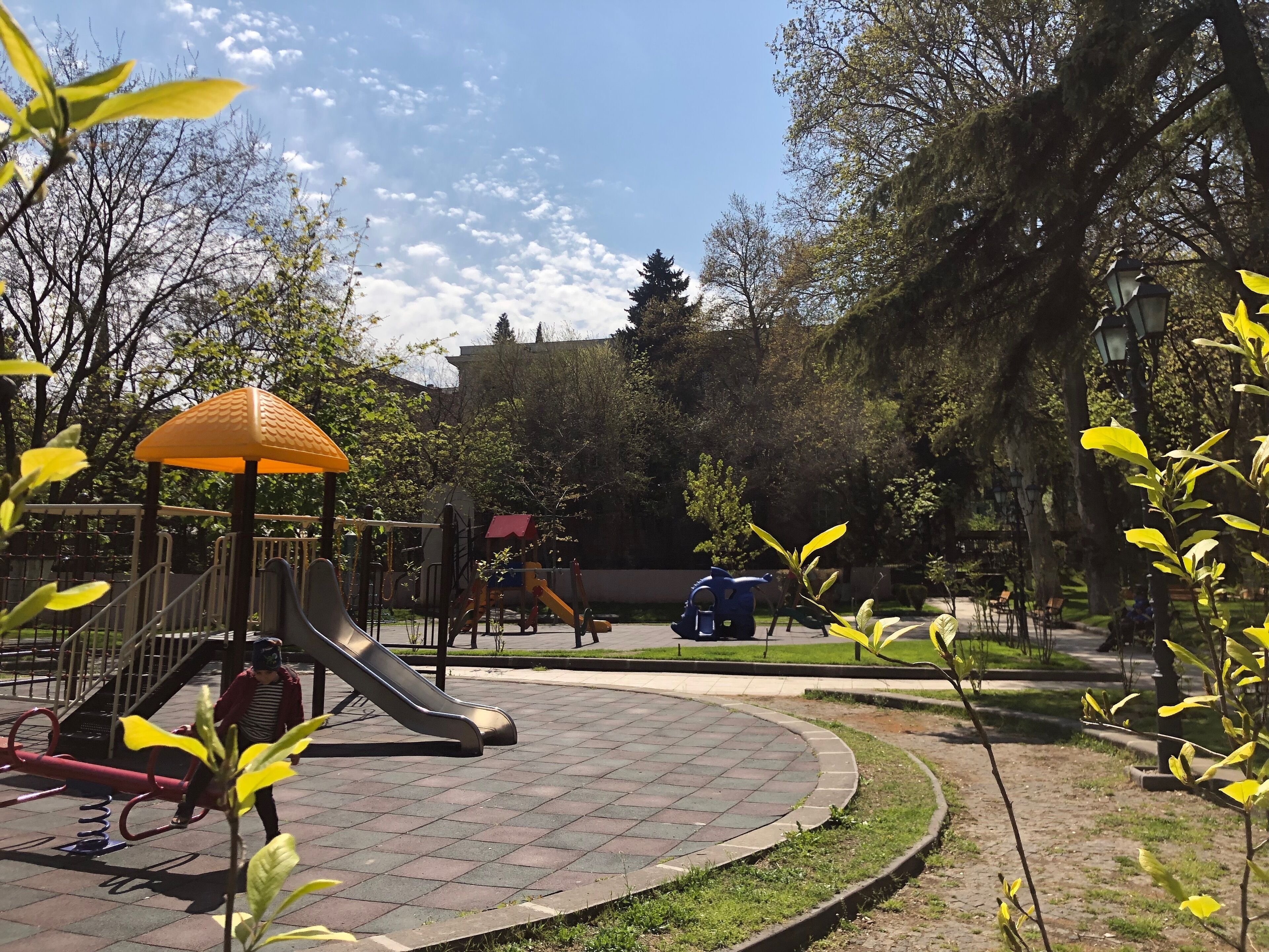 A playground with a slide and climbing structures, surrounded by trees and a paved area.