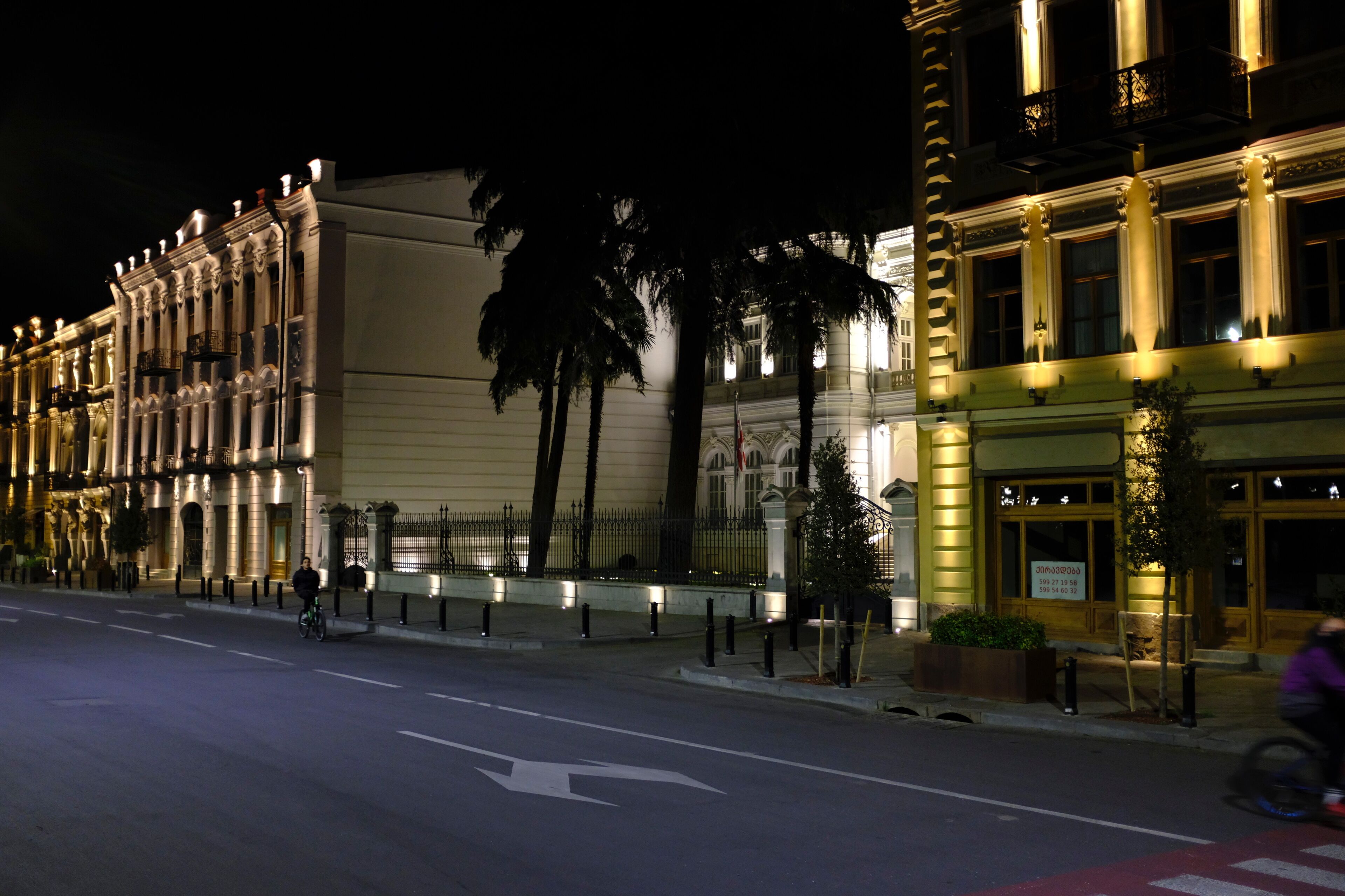 A nighttime street view with a building featuring classical architecture, a cyclist, and a pedestrian.