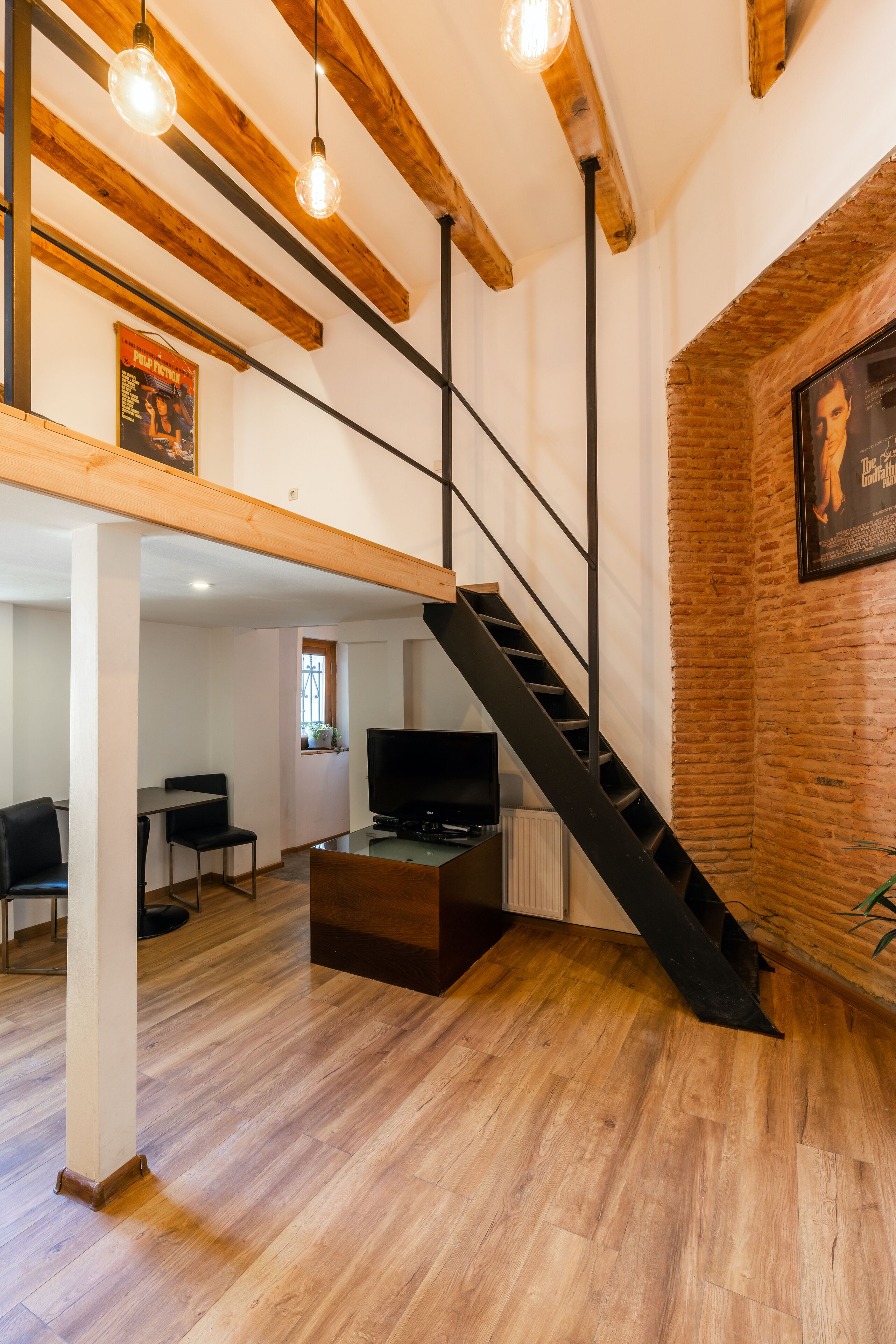 Una sala de estar moderna con vigas de madera a la vista, una escalera negra, un televisor y una pared de ladrillo.