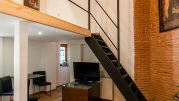 Una sala de estar moderna con vigas de madera a la vista, una escalera negra, un televisor y una pared de ladrillo.