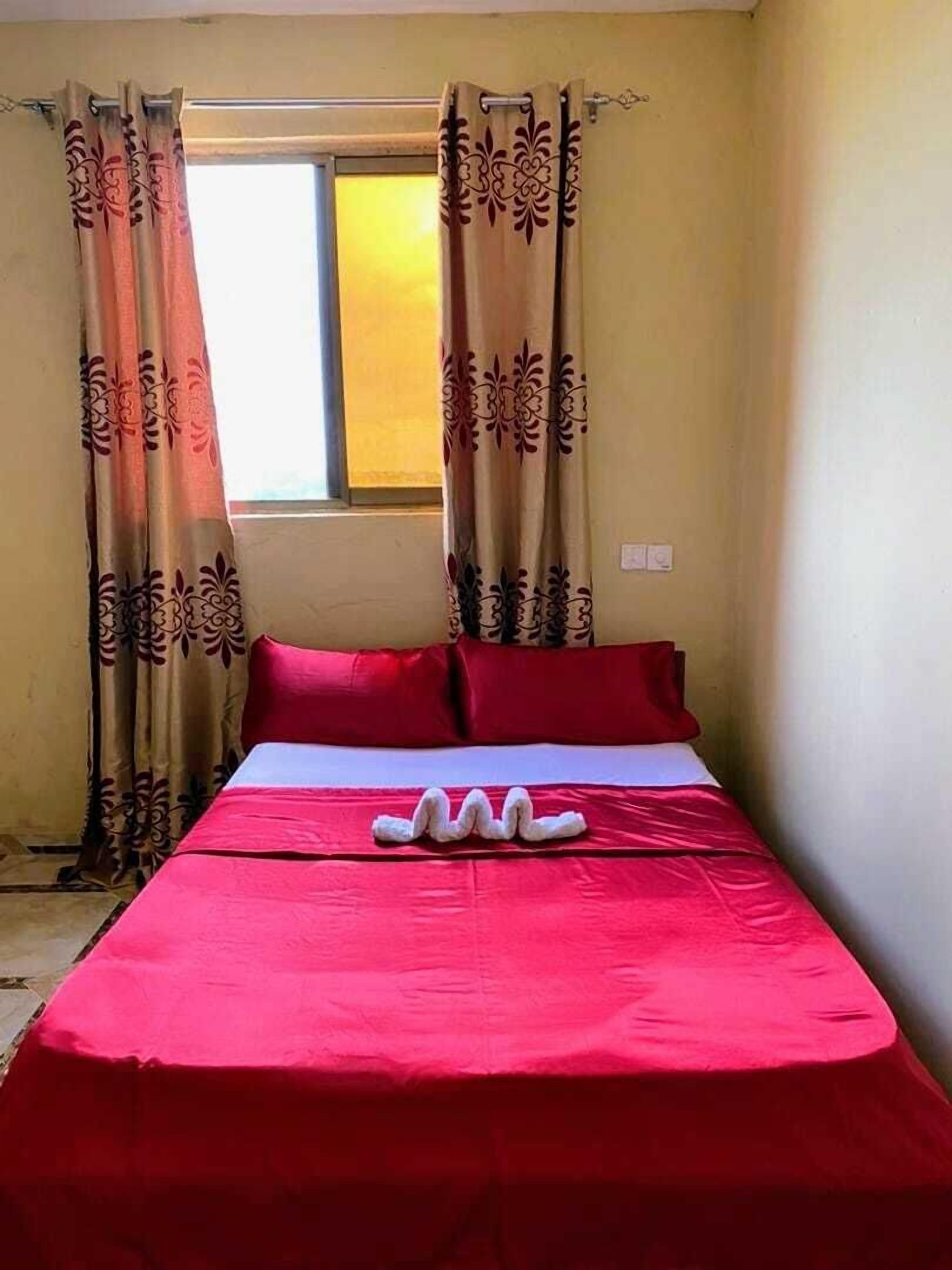 Una cama individual con colcha y almohadas rojas, cortinas con estampado floral y una ventana con vistas al exterior.