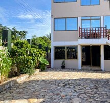 Edifício com área de recepção, entrada com calçada de pedra e vegetação ao redor.