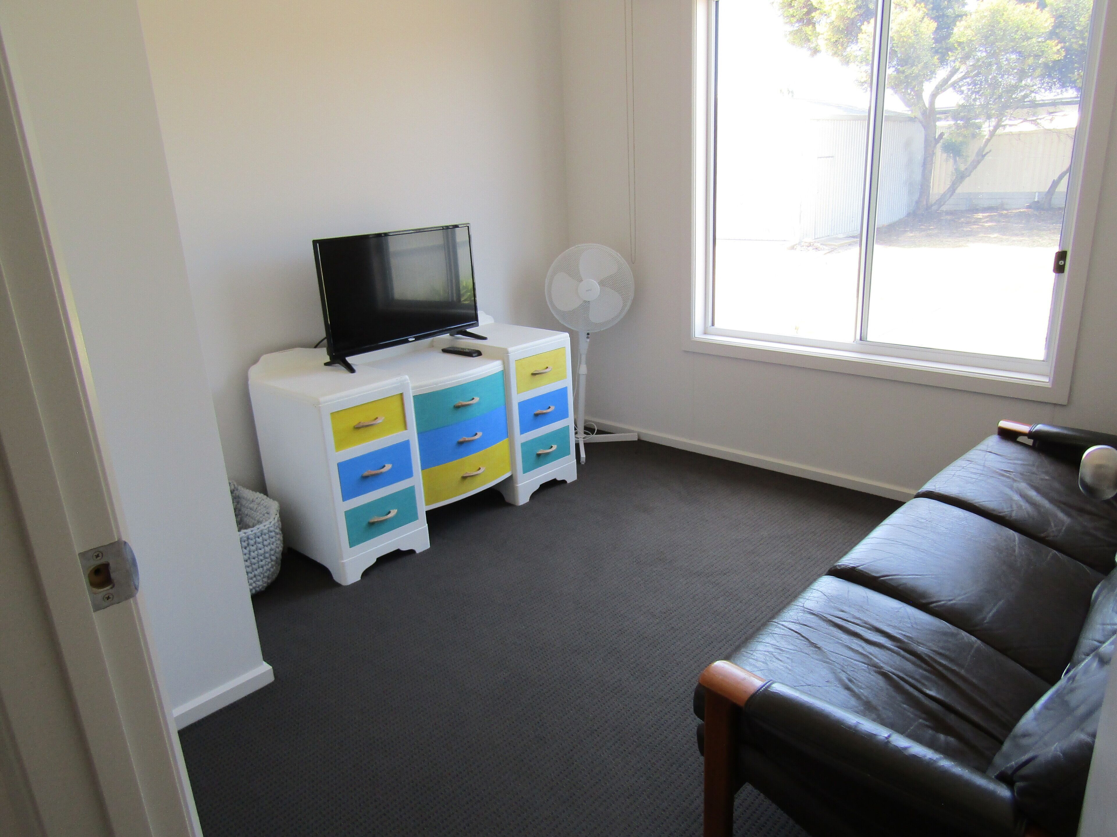 A room with a TV on a white cabinet, a fan, a leather couch, and a window with a view of trees.