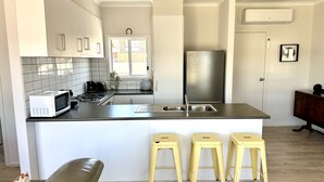 A modern kitchen with white cabinets, a stainless steel refrigerator, a microwave, and a sink. There are three yellow stools and a brown couch.