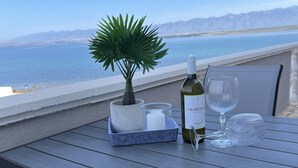 Une table dressée avec une bouteille de vin, un verre, une bougie et une plante, sur un balcon offrant une vue sur la mer et les montagnes.