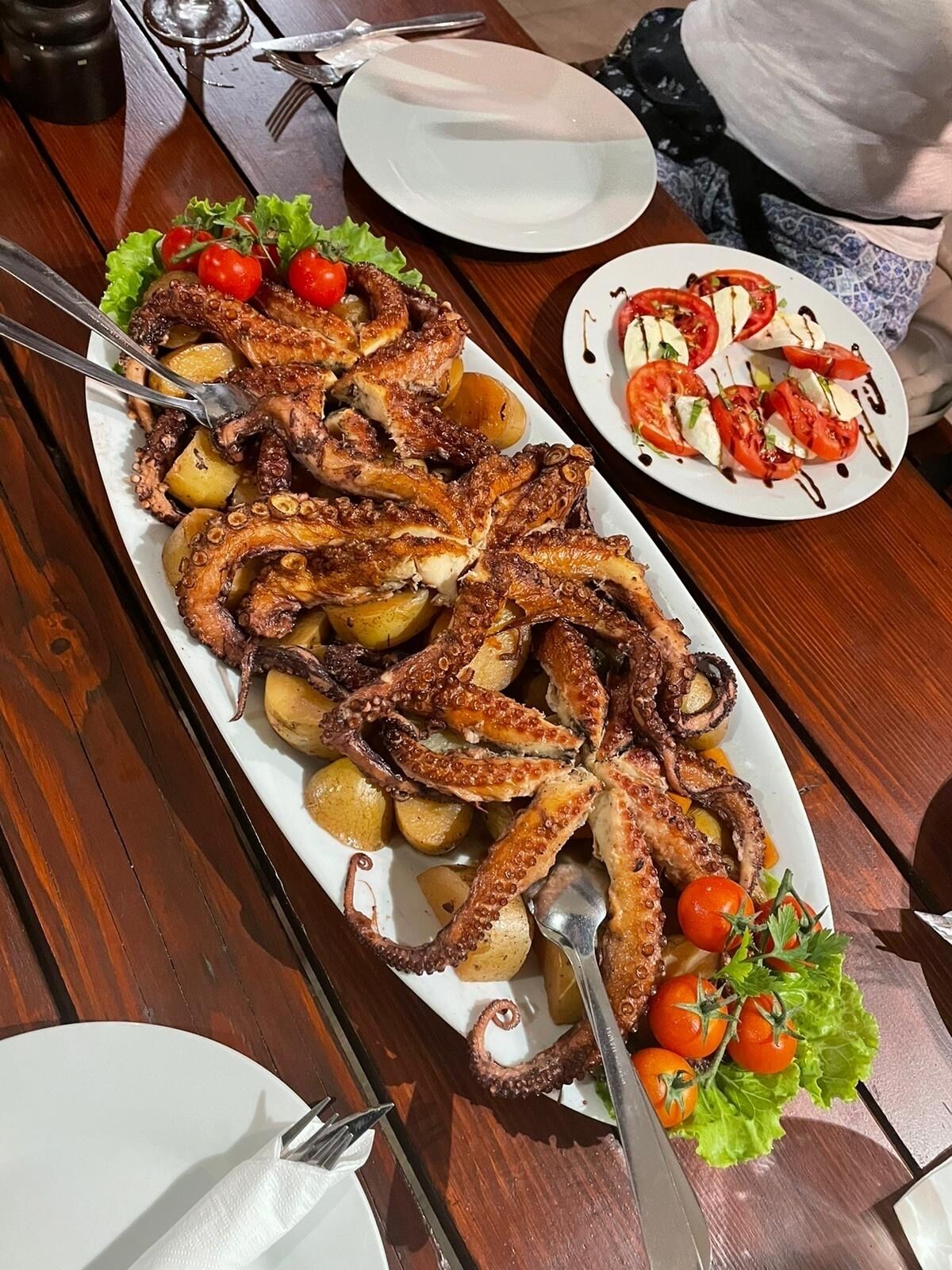 Ein Holztisch mit einem großen Teller gegrilltem Oktopus und Kartoffeln, einem kleineren Teller Tomatensalat und einer Tasse Kaffee.