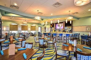 A hotel lobby with multiple dining tables, chairs, a bar area, and a television screen.