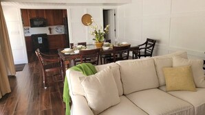 A dining area with a wooden table, chairs, and a white sofa. A kitchen is visible in the background with wooden cabinets and a microwave. - Family friendly condo located in Pineapple Village. (St Thomas)