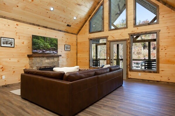 Un intérieur en bois agrémenté d’un grand canapé d’angle, d’une cheminée en pierre et de grandes fenêtres offrant une vue sur les arbres.