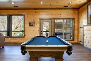 Une table de billard se trouve dans une pièce en bois, dotée de grandes fenêtres et d’une porte donnant sur l’extérieur.