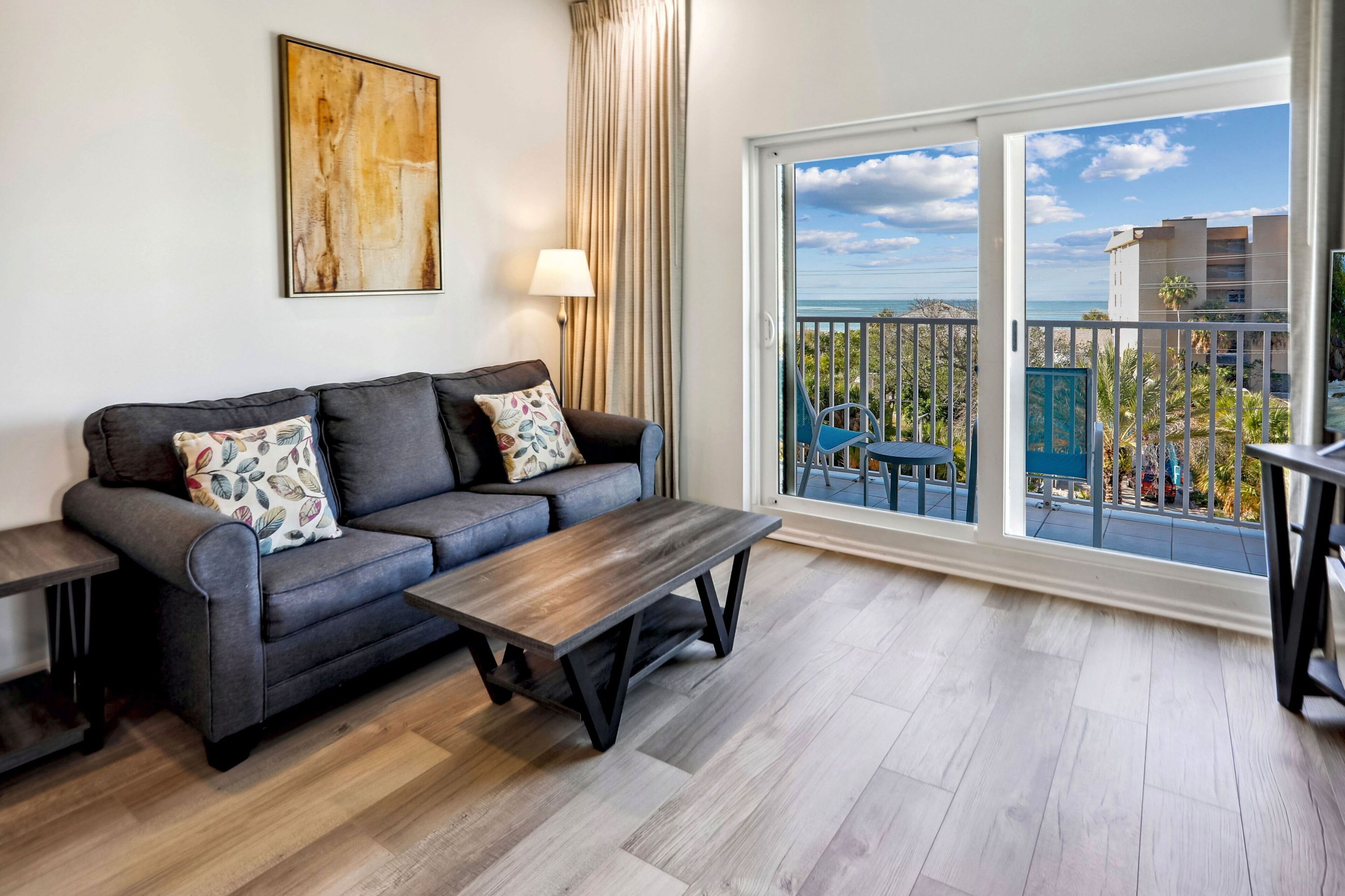 A living room with a sofa, a coffee table, and a large window with a view of the sea and buildings.