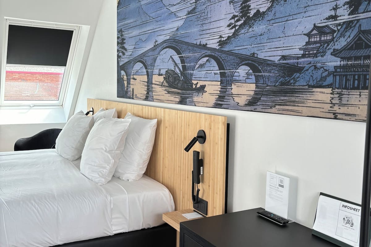 A modern hotel room with a large bed, a wooden headboard, a desk with a computer, and a wall-mounted phone.