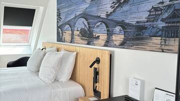 A modern hotel room with a large bed, a wooden headboard, a desk with a computer, and a wall-mounted phone.