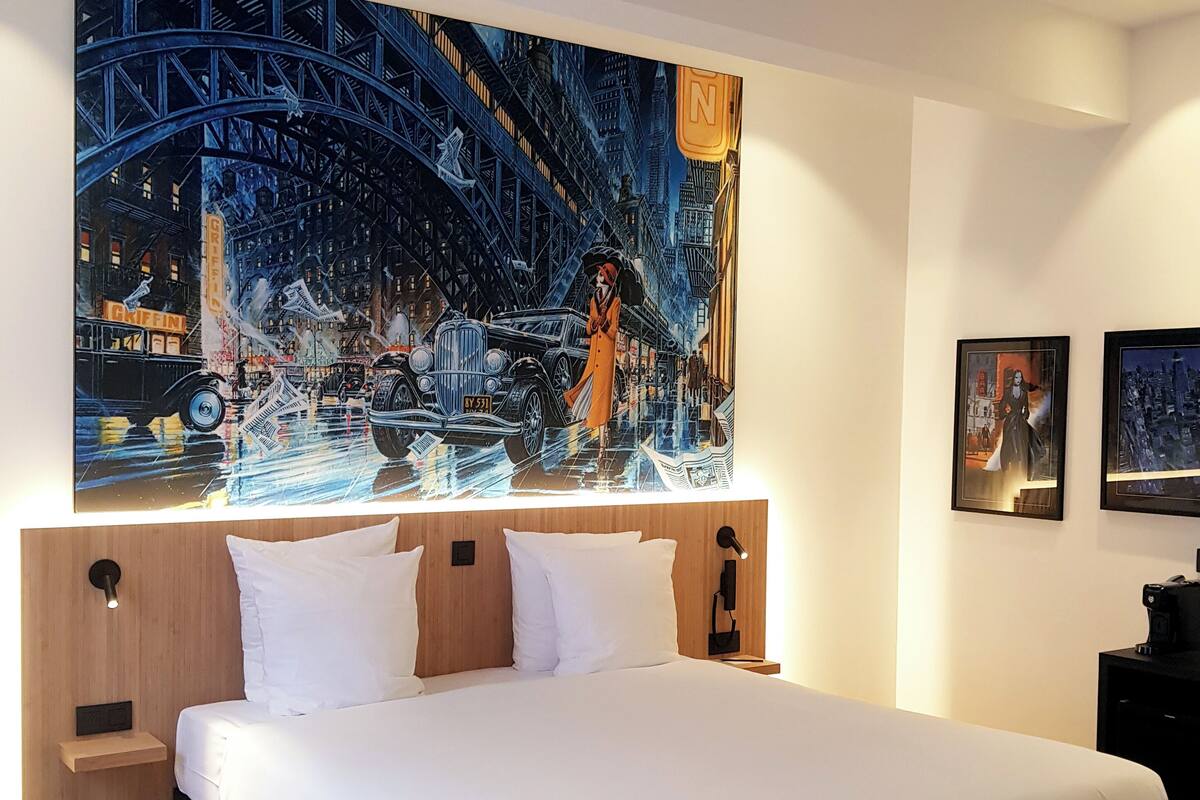 A modern hotel room with a large bed, wooden headboard, and a cityscape painting above it.