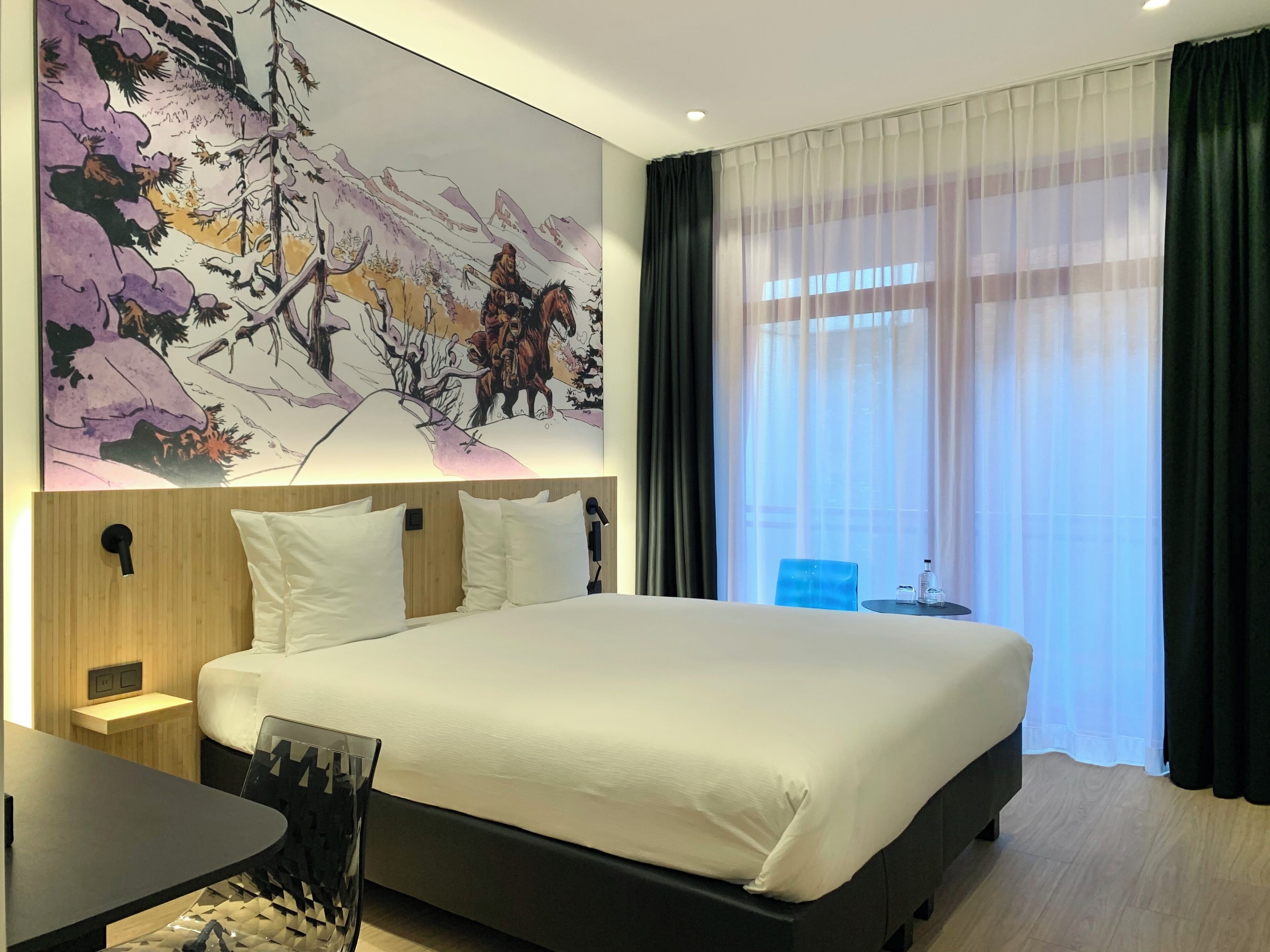 A modern hotel room with a large bed, a desk, and a painting of a mountainous landscape.