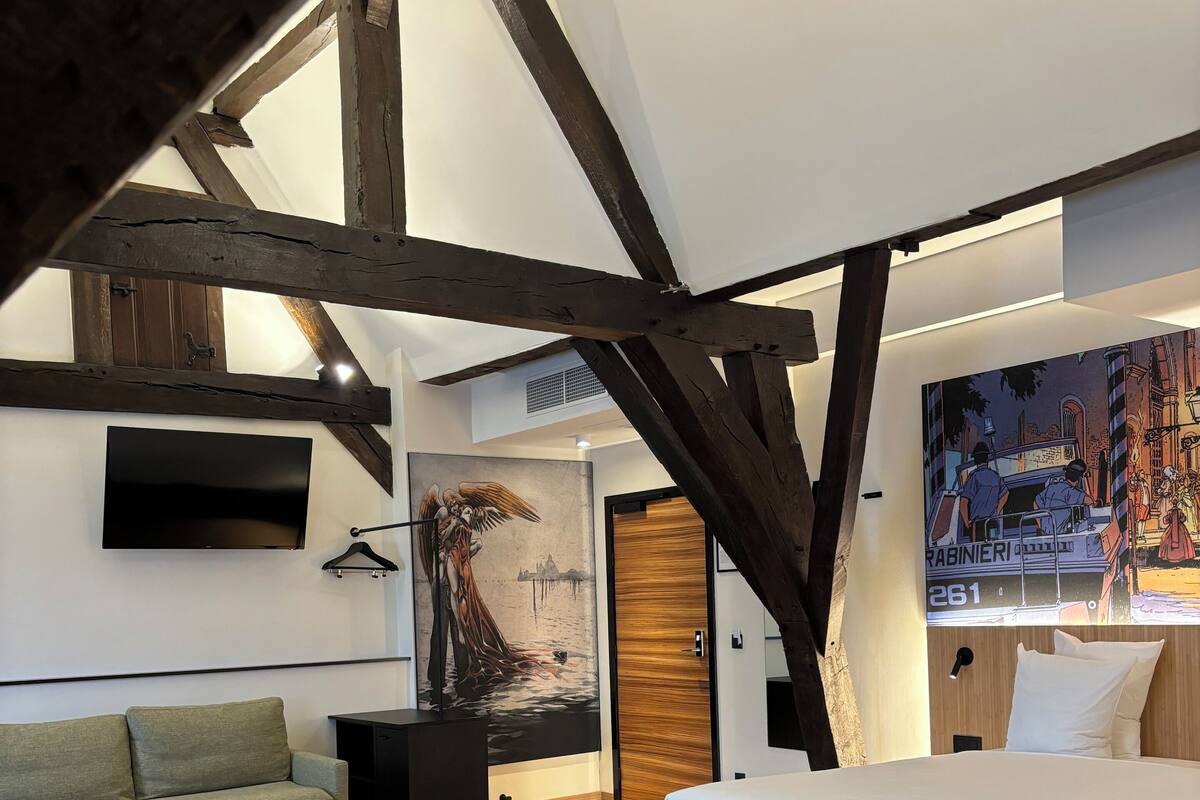 A modern hotel room with a loft-style ceiling, a large bed, a sofa, two green armchairs, a small table, and a painting on the wall.