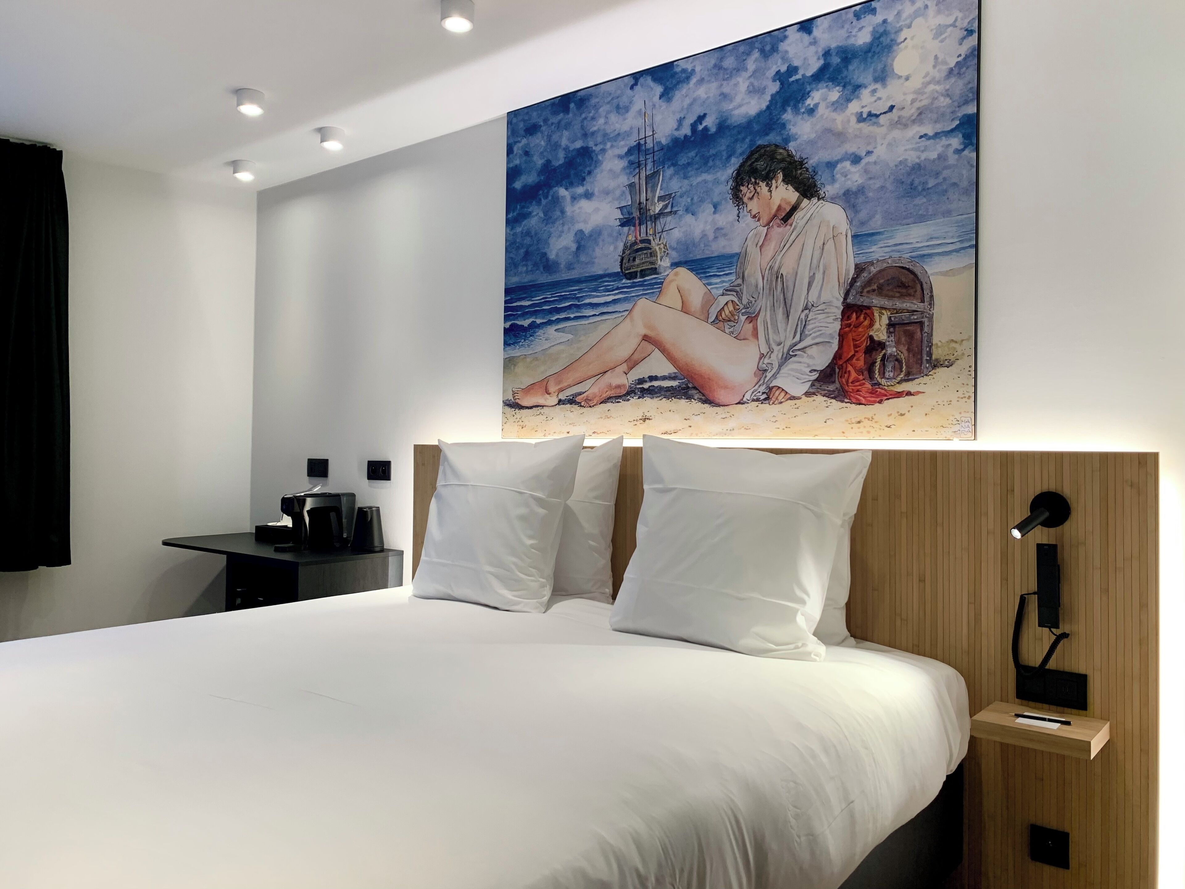 A modern bedroom with a large bed, white pillows, a nightstand, a coffee maker, and a painting of a beach scene.