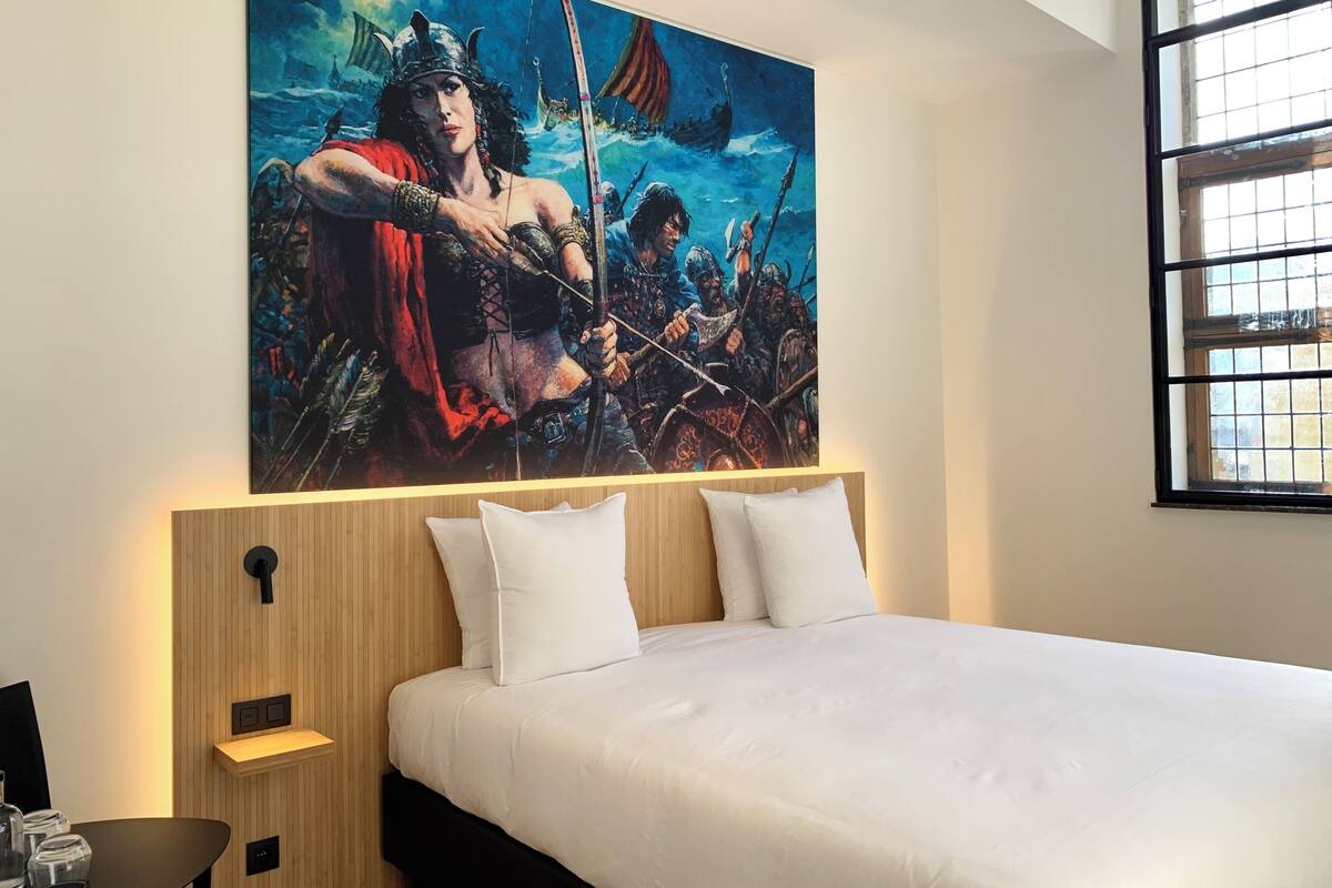 A bedroom with a large bed, two pillows, a wooden headboard, a nightstand, and a painting of a warrior on the wall.