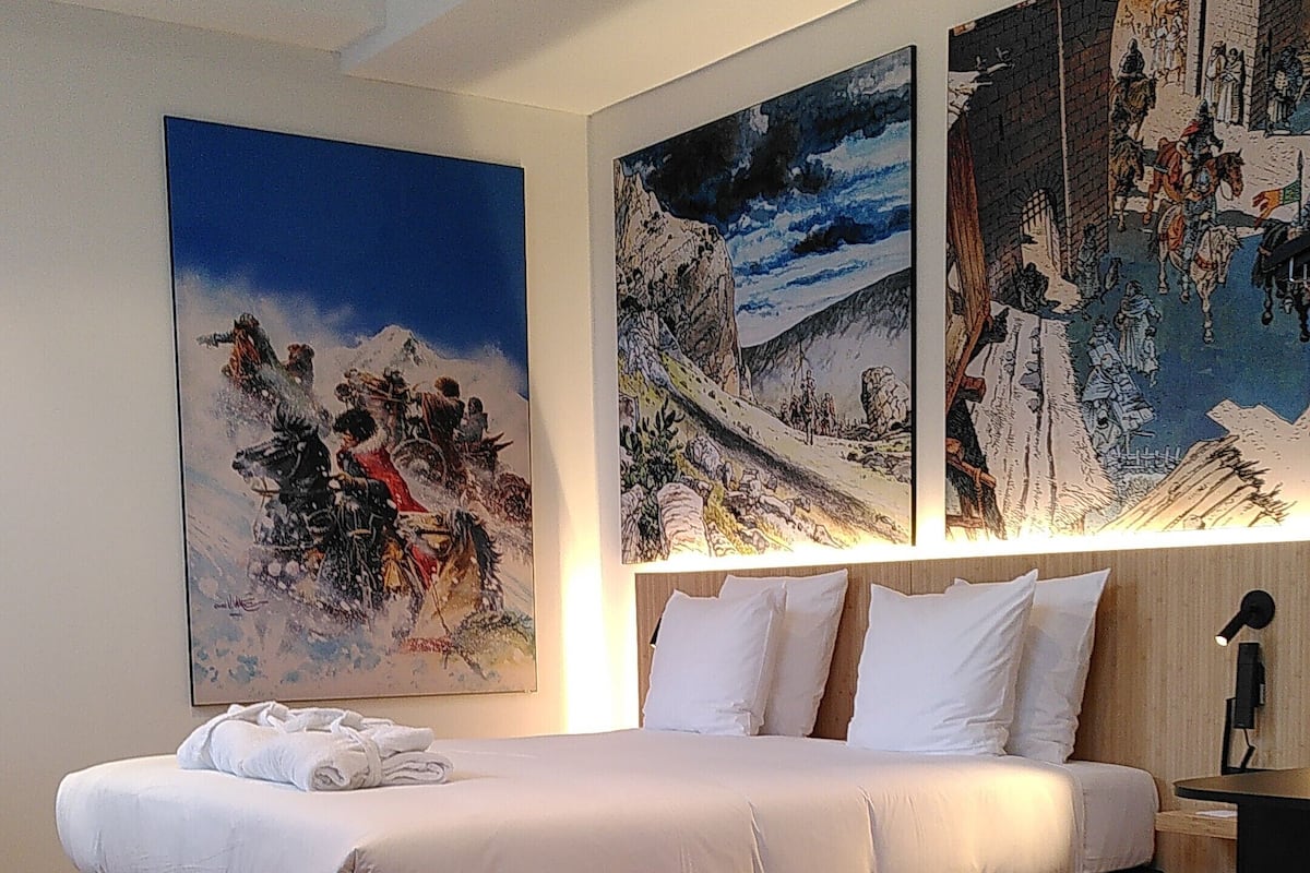 A modern bedroom with a large bed, a green armchair, and two large paintings on the wall.