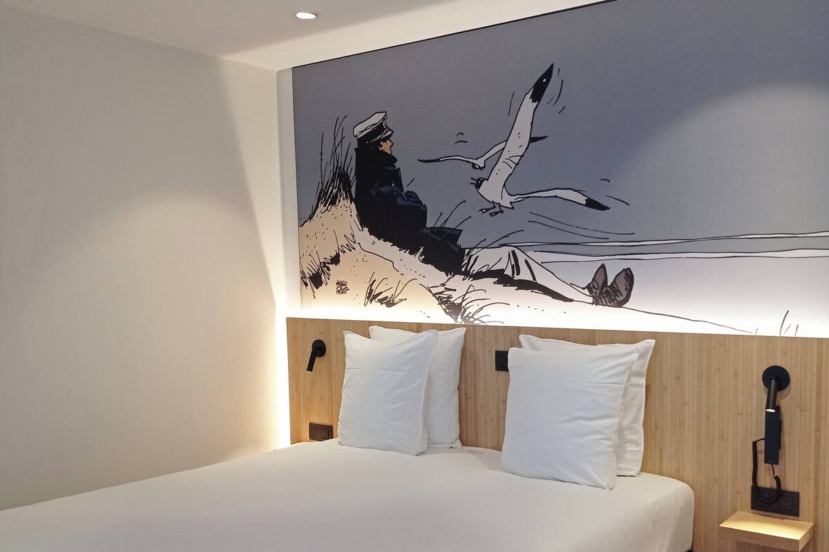 A modern hotel room with a large bed, white bedding, a wooden headboard, and a wall-mounted artwork depicting a seascape.