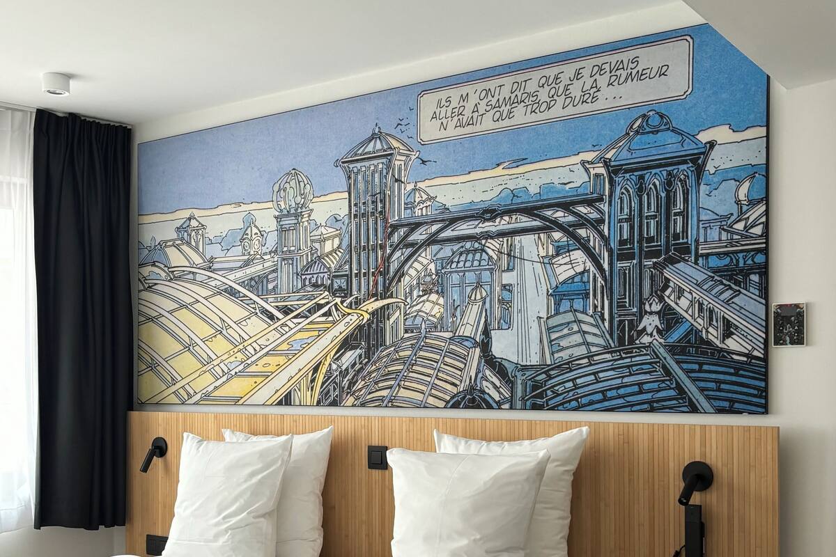 A hotel room with a large bed, white bedding, a wooden headboard, and a cityscape mural on the wall.