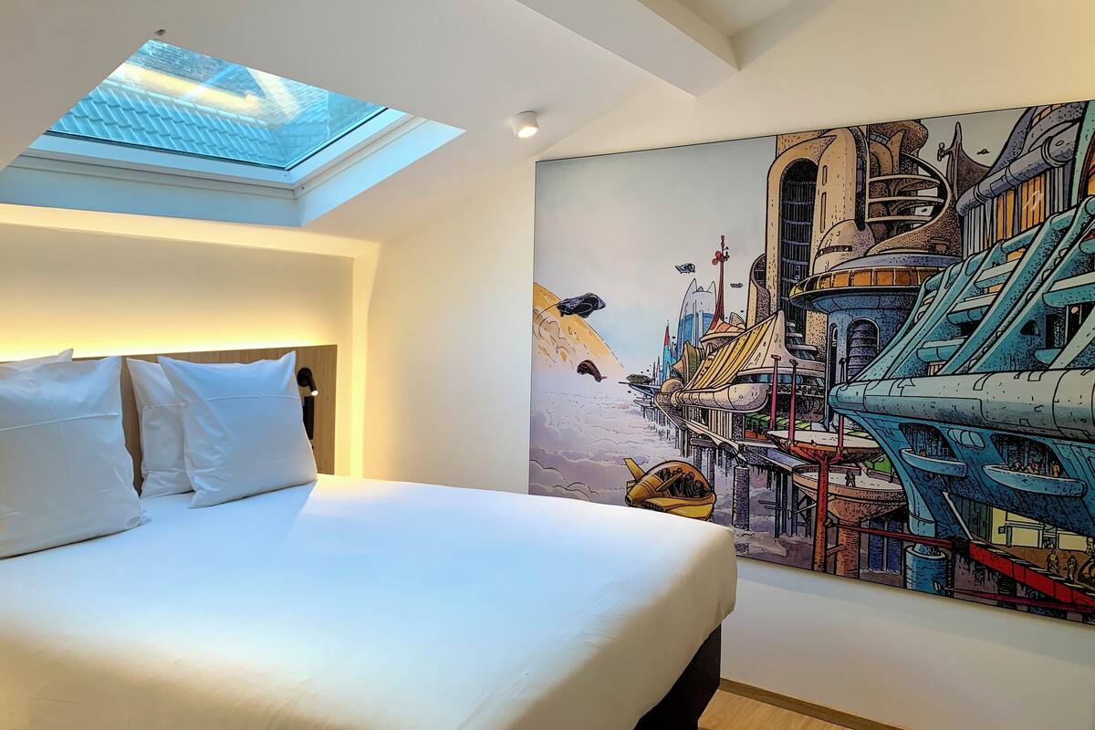 A modern hotel room with a skylight, a large bed with white linens, and a vibrant cityscape mural on the wall.