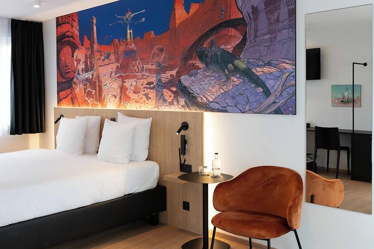 A modern hotel room with a large bed, a desk, a chair, and a large painting on the wall.