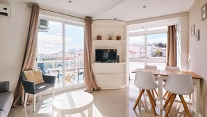 Ein modernes Wohnzimmer mit Flachbildfernseher, Essbereich mit weißen Stühlen und großen Fenstern mit Blick auf die Stadt.