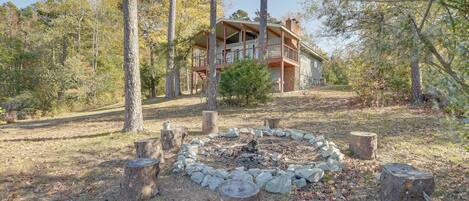 A cabin with a deck, surrounded by trees and a fire pit.