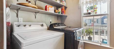 A laundry area with a white washing machine and a black dryer, a shelf with cleaning supplies, and a window with a view of the street.
