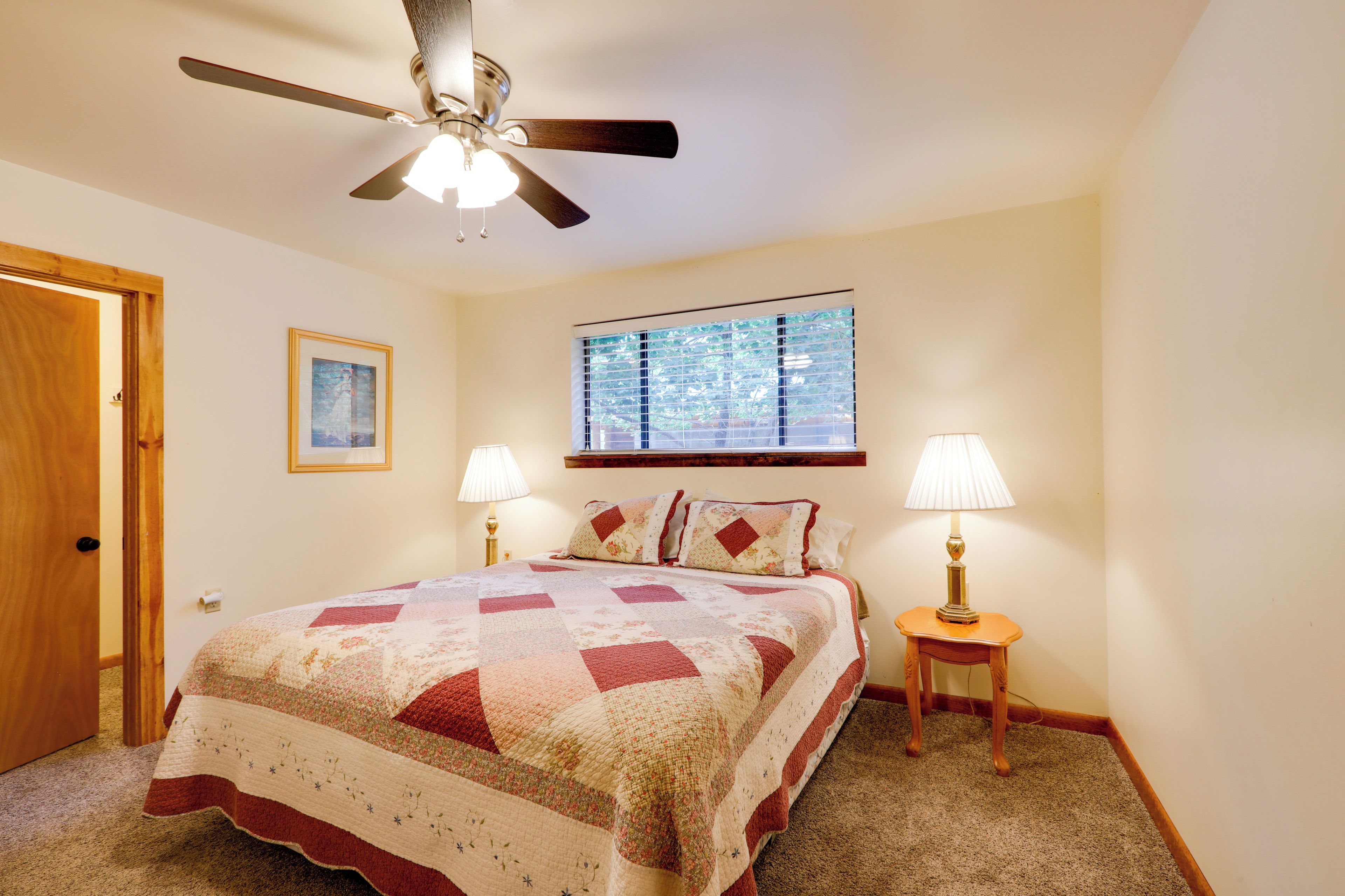 A bedroom with a bed, two bedside lamps, a ceiling fan, a window with blinds, and a door.