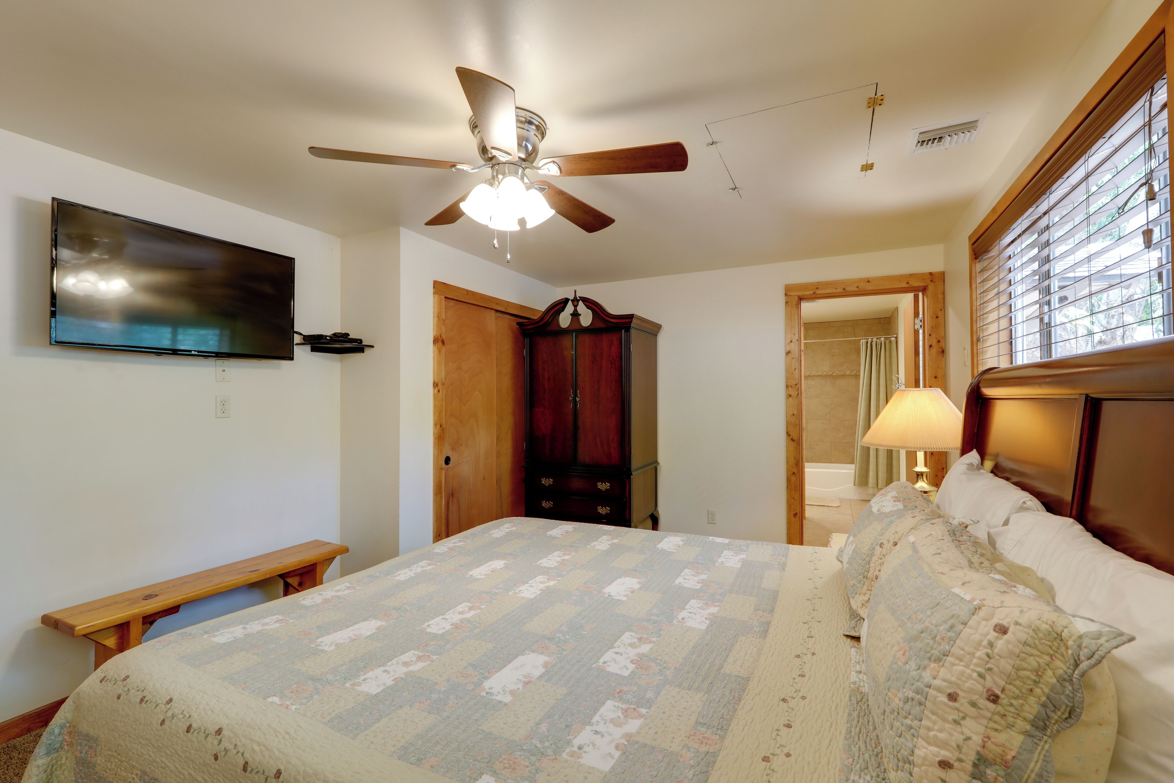 A bedroom with a bed, a ceiling fan, a TV, a wooden wardrobe, and a window with blinds.