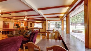Uma sala ampla com uma mesa de jantar de madeira, assentos confortáveis, uma cozinha ao fundo e janelas grandes com vista para o exterior.