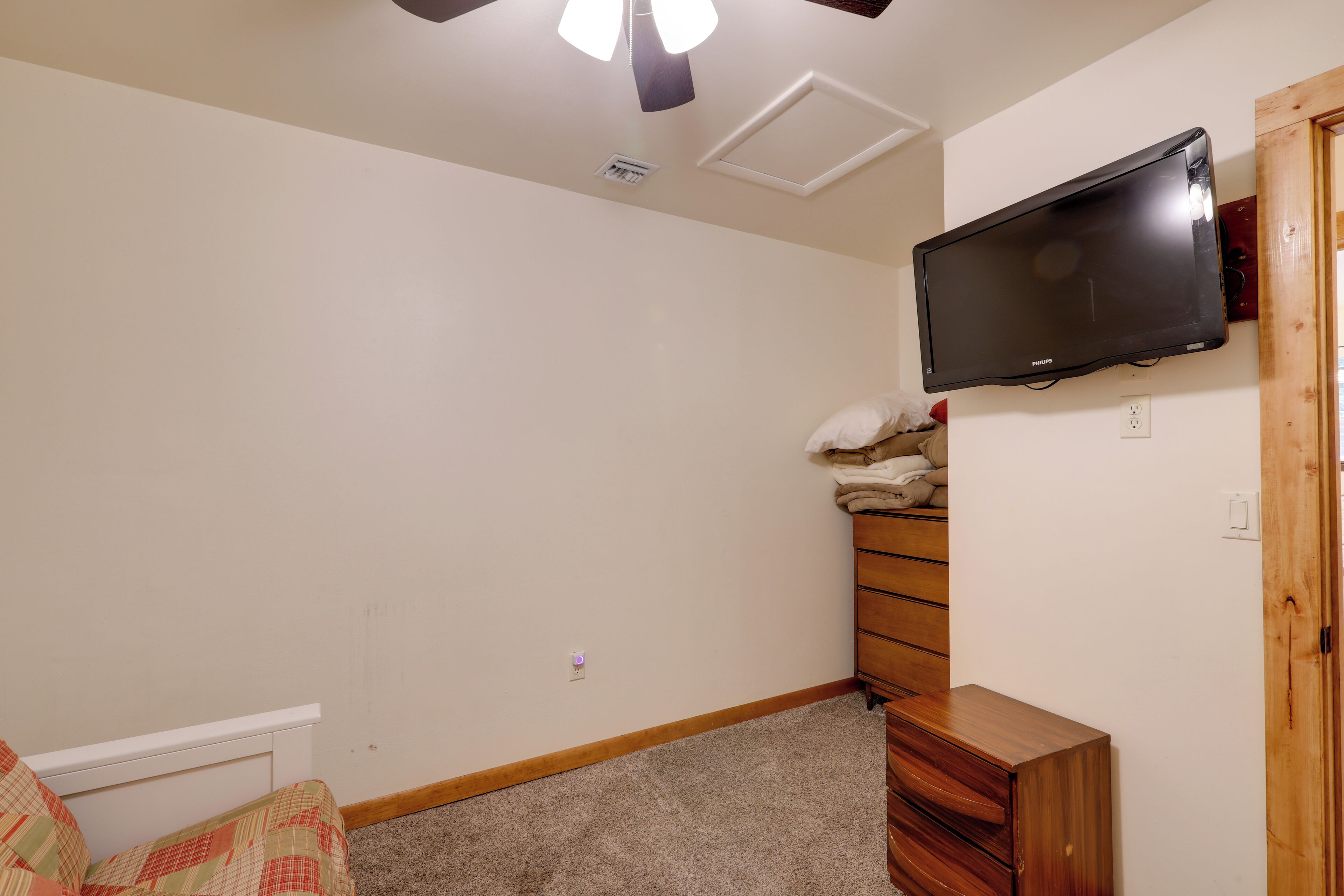 A bedroom with a bed, a dresser, a TV mounted on the wall, and a ceiling fan.
