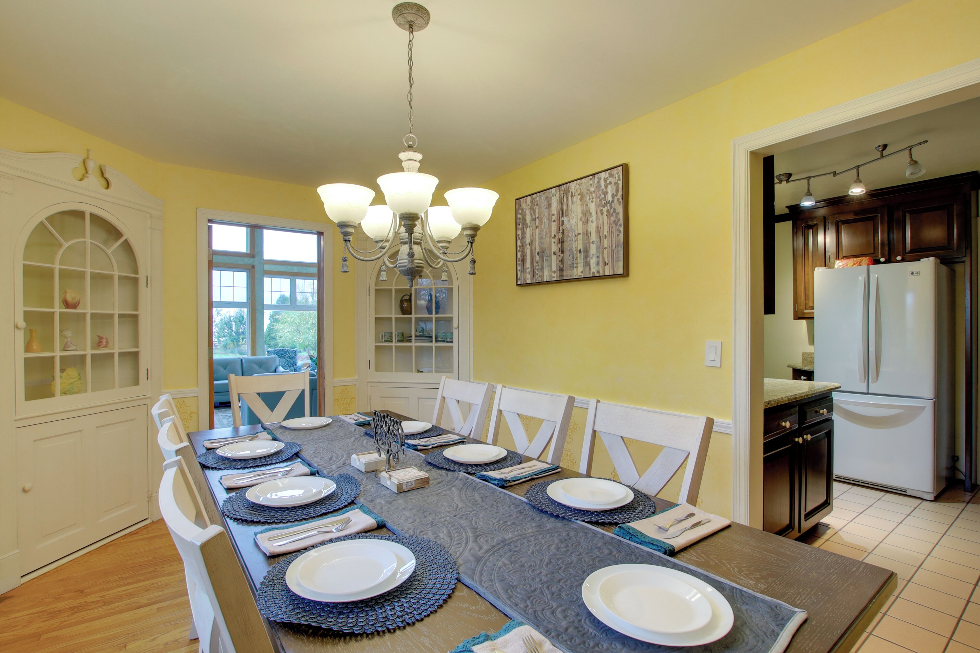 A dining room with a long table set for six, white chairs, a chandelier, and a kitchen visible through an open doorway.